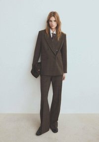 Kobieta w brązowym, podwójnie zapiętym garniturze z rozszerzanymi spodniami, białej koszuli i krawacie, trzymająca pasującą czapkę w pobliżu jasnej, jednolitej ściany.