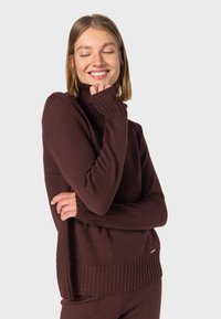 Burgunderfarbener Rollkragenpullover mit gerippten Bündchen und Saum, aus strukturiertem Strickmaterial und mit lockerer Passform. Minimalistisches goldfarbenes Akzentdetail.