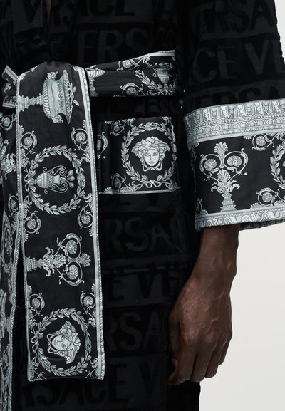 Μαύρη ρόμπα με μαύρο σε μαύρο κείμενο Versace, λευκές περίτεχνες floral και βάζο εκτυπώσεις, έμβλημα κεφαλής Μέδουσας στη ζώνη και στο μανίκι, εμφανίζεται σε ένα χέρι.