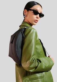 Zaino in tessuto nero con design curvo, dotato di due compartimenti con zip. Indossato sopra una giacca di pelle verde, che mette in risalto la texture elegante della borsa.