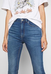 Sinisiä farkkuhousuja, joissa on keskitason pesu, perinteinen viiden taskun muotoilu ja istuva siluetti, yhdistettynä graafiseen valkoiseen t-paitaan.