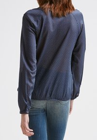 Námořnická modrá blůza s bílými puntíky, dlouhými rukávy, elastickým lemem a knoflíkovými detaily na manžetách. Hladká tkanina a volný střih.