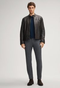 Schwarze Lederjacke, marineblauer Pullover und strukturierte graue Hose. Model trägt braune Wildlederstiefel. Unifarbene Töne und taillierte Passform.