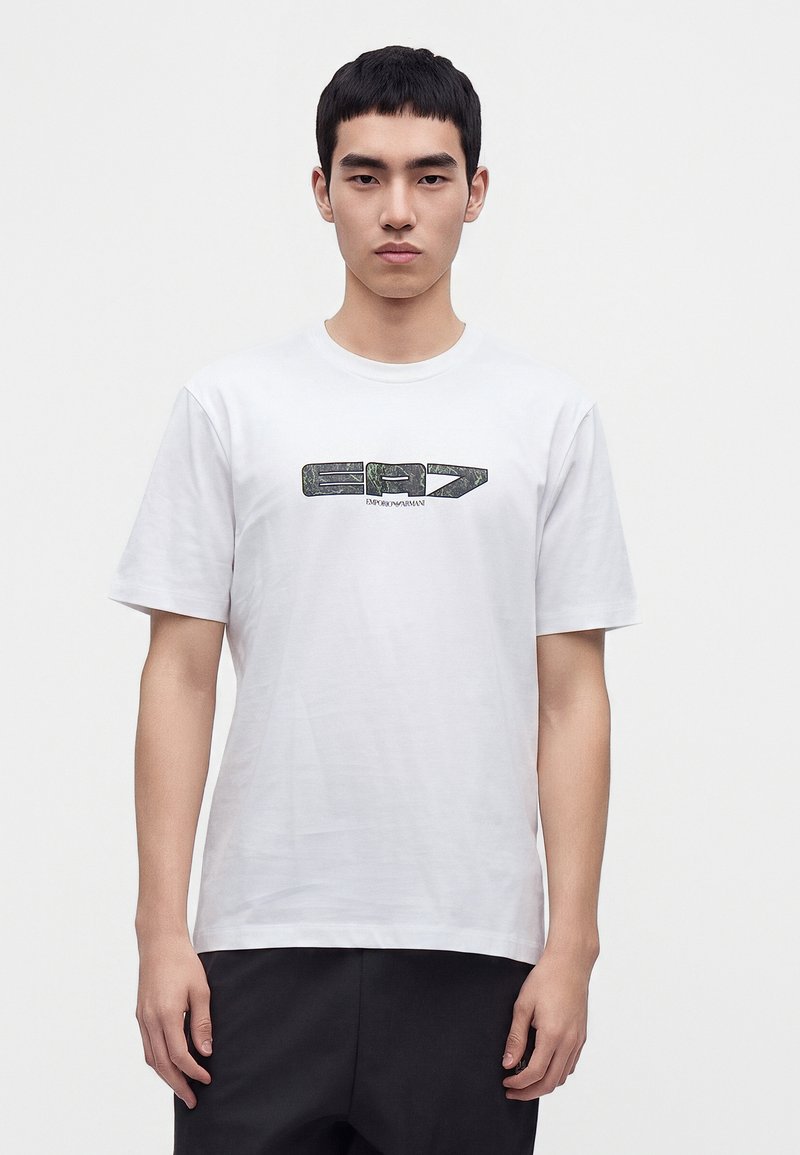 Νέος άνδρας που φοράει λευκό T-shirt με μαύρο γραφικό λογότυπο και μαύρο παντελόνι, στέκεται μπροστά σε ένα απλό φωτεινό φόντο.