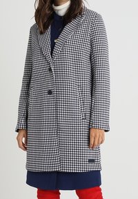 Žena v černobílé kostkované vrstvené kabátu po kolena s knoflíky, námořnickém šatu, bílém roláku a červených kalhotách, stojící na bílém pozadí.