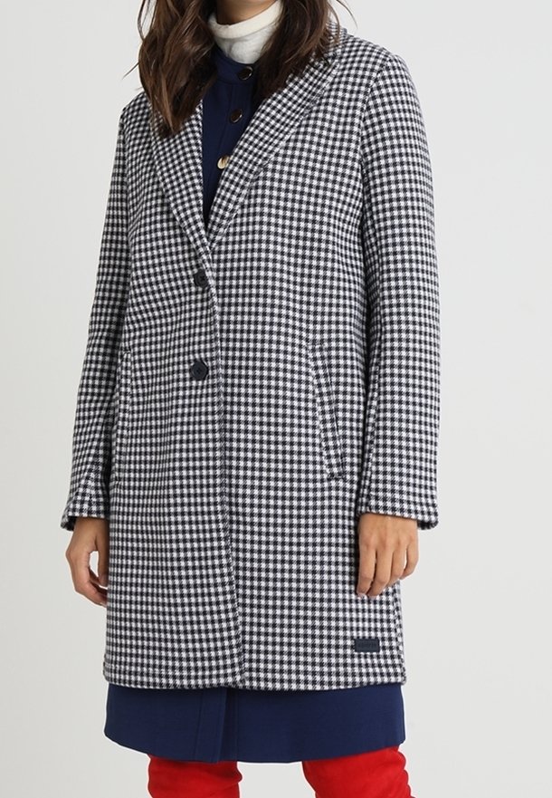 Žena v černobílé kostkované vrstvené kabátu po kolena s knoflíky, námořnickém šatu, bílém roláku a červených kalhotách, stojící na bílém pozadí.