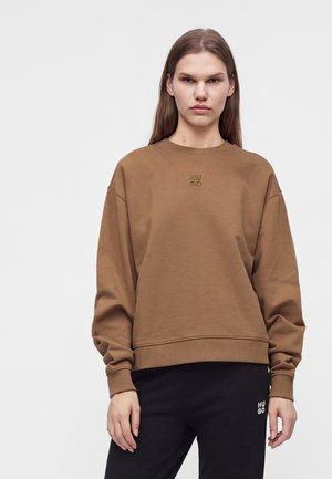 Kvinde med glat brunt hår iført en brun sweatshirt og sorte bukser, begge med små "Hugo" logoer.