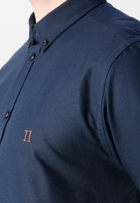 Mörkblå button-up skjorta i texturerad tyg med krage, två knappar vid halsen och en liten orange "II" brodering på bröstet.