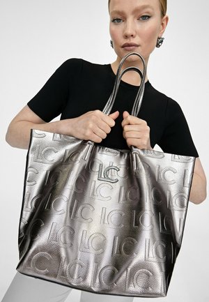 Metalická stříbrná taška přes rameno s embosovaným vzorem loga, dvojitými uchy a výrazným kovovým akcentem. Hladký povrch s pevným tvarem.