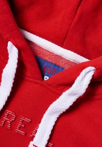 Rode hoodie met contrasterende witte trekkoorden, voorzien van een zachte binnenkant en een label met "SUPERDRY" in het blauw. Gestructureerde stof en logodetail.