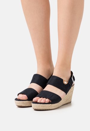 Sandales à talons compensés en suede noir avec deux larges straps, semelle en jute tressé, bout ouvert et bride de cheville réglable avec un accent métallique.