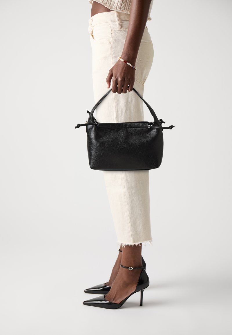 Black leather handbag with a rounded top handle, rectangular shape, subtle texture. Worn with light-colored, cropped jeans and black stilettos.
