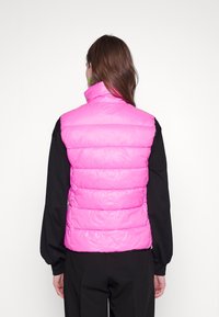 Frau mit einer leuchtend pinken Daunenweste über einem schwarzen Langarmshirt, die sich abkehrt, mit langen braunen Haaren und grünen Creolen.