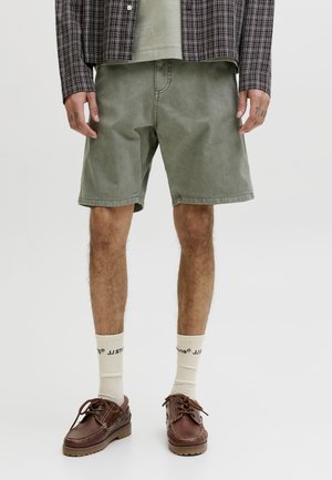 Άνδρας που φορά χακί σορτς, κρεμ κάλτσες με μαύρο κείμενο, καφέ δερμάτινα παπούτσια, καρό πουκάμισο και πράσινο μπλουζάκι ορατό κάτω από το τελείωμα του πουκαμίσου.