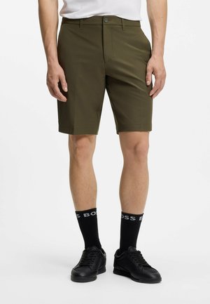 Mand iført olivengrønne shorts til knæet, sorte sokker med "BOSS"-bogstaver og sorte sneakers, der står mod en hvid baggrund.
