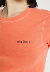 Close-up of person wearing orange crew neck t-shirt with small black embroidered text "iets trans..." on the left chest.