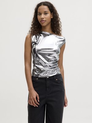 Kvinne med krøllete hår iført skinnende sølvfarget ermeløs topp og høytlivs svarte bukser, stående foran en ensfarget lys bakgrunn.