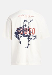 Kreminės spalvos medvilninis marškinėlių su grafika, vaizduojančia rodeo kovotoją ant šokinėjančio žirgo, su raudonais ir tamsiai mėlynais teksto akcentais ant nugaros.