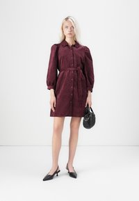Bordeauxfarbene Samt-Wickelkleid mit Puffärmeln, Taillengürtel, Knopfverschluss und leicht ausgestellter A-Linie, kombiniert mit schwarzen Slingback-Absätzen.