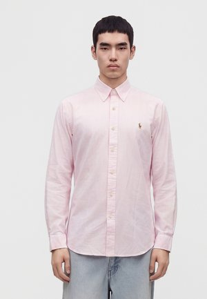 Jeune homme debout portant une chemise à rayures rose clair à boutons et un jean bleu clair sur un fond blanc uni.