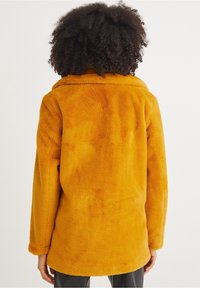 Cappotto giallo senape, morbido e peloso, con colletto e maniche lunghe. Texture morbida con vestibilità ampia, caratterizzato da una schiena liscia e dritta.