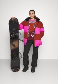Doudoune brune, rose et noire avec un col haut, associée à un pull à motifs, un pantalon noir et des bottes de neige, tenant un snowboard.
