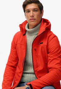 Veste extérieure rouge avec capuche, dotée d'une poche zippée et d'accents noirs. Superposée dessous, un pull gris clair côtelé.