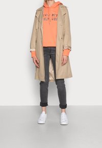 Beige trench coat päällä oranssia hupparia, jossa on painettu teksti, harmaiden farkkujen ja valkoisten tennareiden kanssa. Silkkinen kangas ja käännetyt hihansuut.