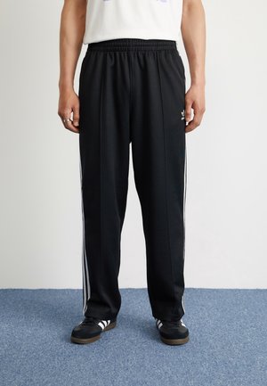 Person står iført sorte Adidas træningsbukser med hvide sidelister, sorte Adidas sneakers og en hvid skjorte foran en hvid væg og blåt tæppe.