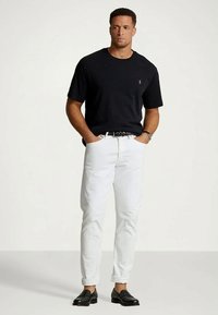 Homme portant un t-shirt noir à manches courtes, un pantalon blanc retroussé, une ceinture noire, une montre noire et des mocassins noirs, se tenant avec les mains dans les poches.