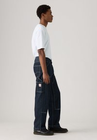 Homem em pé de lado, usando uma t-shirt branca, jeans largos de cor azul escura com bolsos de remendo, e sapatos de couro pretos, num fundo simples.