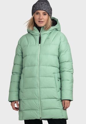 Vrouw die een lichtgroene doorgestikte winterjas met capuchon en een grijze gebreide muts draagt, staand tegen een effen lichte achtergrond.