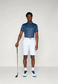 Muž stojí a drží golfovú palicu, oblečený v námorníckej polokošeli, bielych šortkách, bielom opasku, bielych ponožkách a bielych golfových topánkach na jednoliatom pozadí.