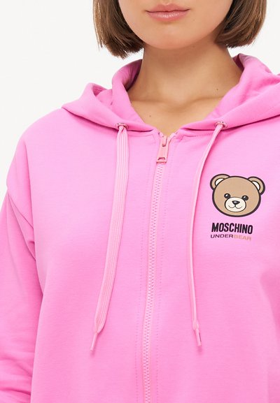 Roza zip-up kapuca iz mehkega materiala, ki vključuje grafiko medveda in besedilo "MOSCHINO UNDERBEAR". Ima tudi vrvice z roza konicami.