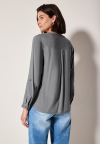 Blouse grise à manches longues en tissu léger et doux, avec un plissé au dos et des poignets boutonnés, associée à un jean en denim bleu clair.