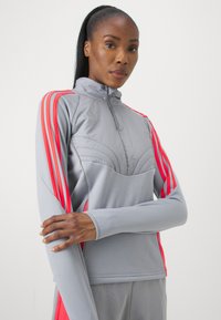 Giacca atletica grigia chiara con colletto alto, caratterizzata da strisce rosa brillante lungo le maniche e elementi di design testurizzati.