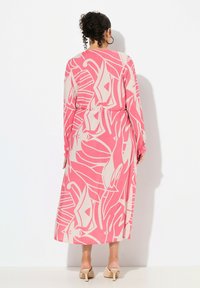 Langes, pinkes Kleid mit abstraktem cremefarbenem Blumenmuster. Leichter Stoff, lockere Passform, elastischer Taillenbund und lange Ärmel. Die Absätze sind hell gefärbt.