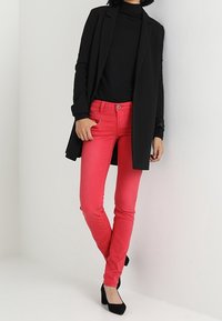 Blazer noir à manches longues porté sur un col roulé noir, associé à un jean slim rose vif et des chaussures noires à talons hauts. Texture de tissu lisse.