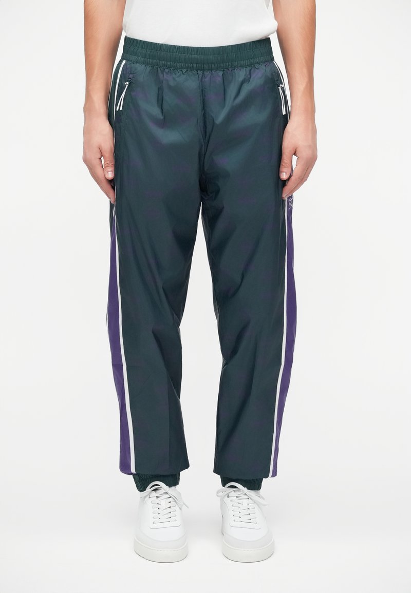 Marineblaue Windbreaker-Hosen mit weißen und lila Seitenstreifen, elastischem Bund und Reißverschlusstaschen. Kombiniert mit weißen Turnschuhen.