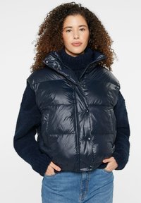 Navy puffer vest with a shiny finish, featuring a high collar, front snap buttons, and patch pockets, layered over a navy sweater.