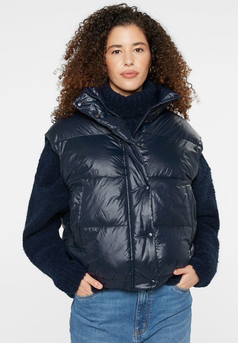 Navy puffer vest with a shiny finish, featuring a high collar, front snap buttons, and patch pockets, layered over a navy sweater.