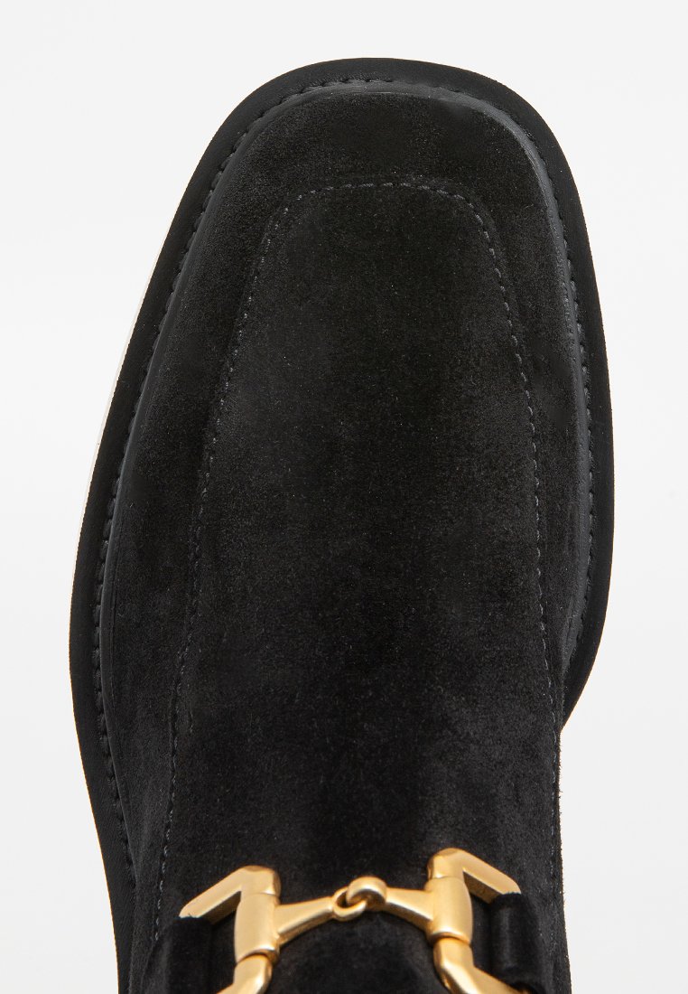Bota de ante negra con punta redonda, que presenta un brillante hebilla metálica dorada y costuras en contraste a lo largo de los bordes.