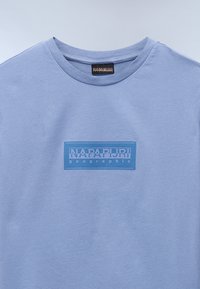 Lyseblå bomulds t-shirt med et rektangulært blåt mærke med hvid tekst "NAPAPIJRI geographic" i midten. Klassisk rund halsudskæring.