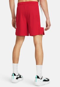 Pantalones cortos de baloncesto rojos con paneles laterales blancos, cintura elástica y aberturas laterales, combinados con zapatillas negras y teal y calcetines blancos.