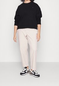 Sudadera negra, pantalones beige claros y zapatillas de deporte en negro y blanco. La sudadera tiene cuello redondo y un corte holgado. Las zapatillas presentan un diseño con patrones.