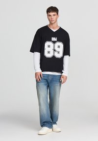 Jersey nera oversized con il numero bianco "89" e dettagli grafici, indossata sopra una maglietta bianca a maniche lunghe; abbinata a jeans azzurri chiari e scarpe bianche.