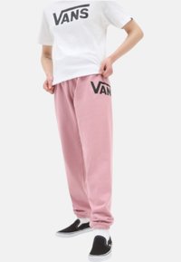 Valkoinen T-paita, jossa on musta "VANS"-logo, yhdistettynä vaaleanpunaisten collegehousujen kanssa, joissa on samanlainen logo, ja mustat liukupohjasandaalit jalassa.
