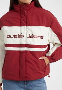 Rød og cremefarvet vindjakke med høj krave, elastiske manchetter, forlommer og "Guess Jeans" logo. Har striber og en glat tekstur.