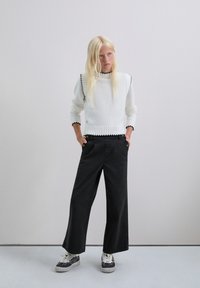 Suéter blanco de punto con terminaciones en negro en forma de concha, combinado con pantalones negros de corte holgado y zapatillas con estampado. Fondo minimalista.