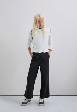 Suéter blanco de punto con terminaciones en negro en forma de concha, combinado con pantalones negros de corte holgado y zapatillas con estampado. Fondo minimalista.